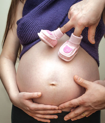 A woman holds her pregnant belly while a man’s hand positions dancing baby shoes on her tummy.